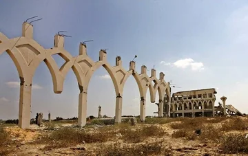 صف طويل من أقواس المطار القائمة يقود نحو مبنى مهدّم تحت سماء زرقاء.