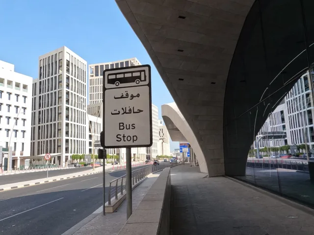 لوحة موقف حافلات ثنائية اللغة على رصيف مشاة بمحاذاة مبانٍ حديثة وطريق للمركبات.